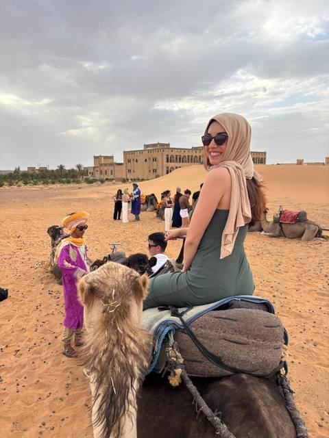       People riding camels in front of a desert building in Morocco.
  