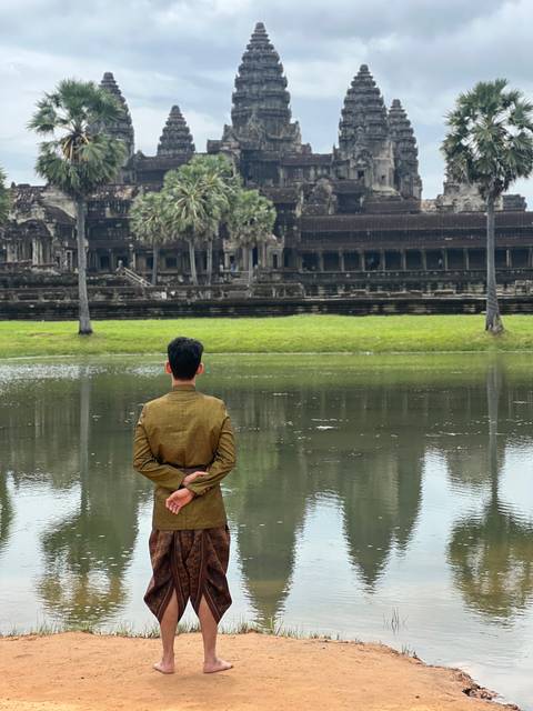      A person with back turned, standing on a grassy area beside a pond, with the iconic Angkor Wat temple in the background.
  