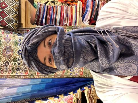 A person wearing a traditional headscarf stands in a shop decorated with colorful textiles.