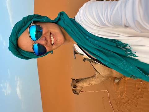 A woman in traditional attire taking a selfie with a camel in the desert.