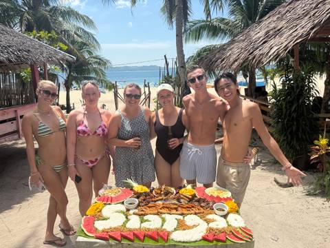      Group of people on a beach enjoying a meal with palm trees in the background.
  
