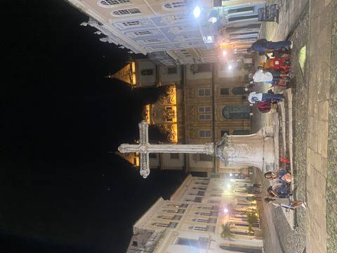 Nighttime scene of a historic building with a cross monument and people mingling.