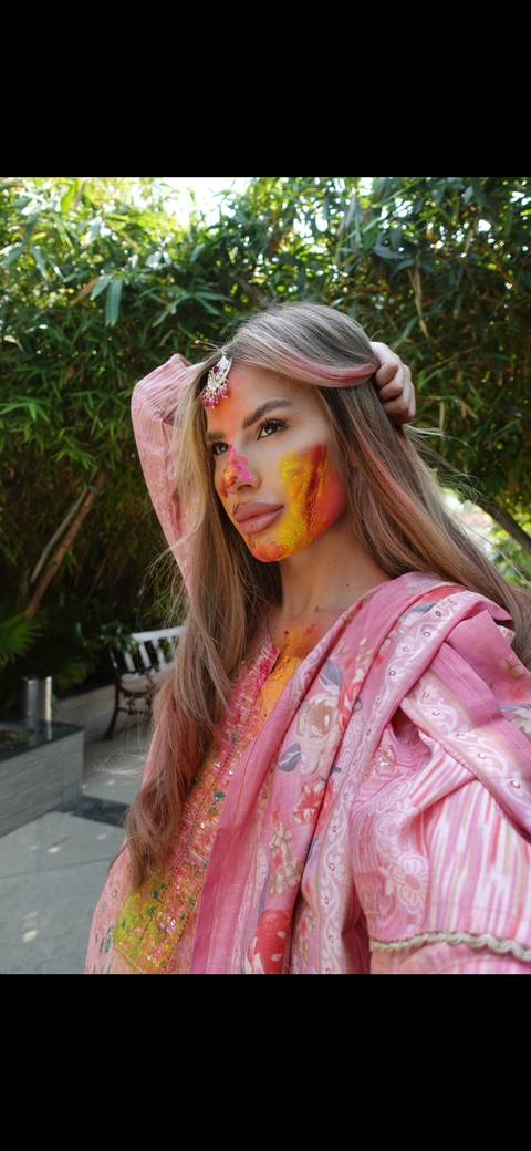 A person with colorful powder on their face, likely during a traditional Indian festival, wearing a pink outfit.