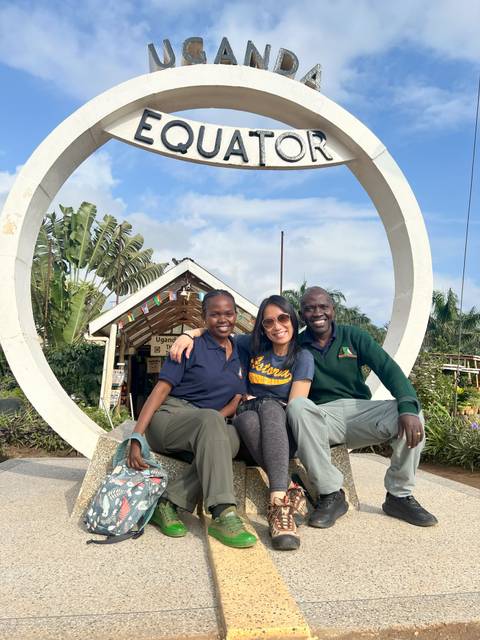 Three people posing at a welcome sign for Uganda by a tropical backdrop.