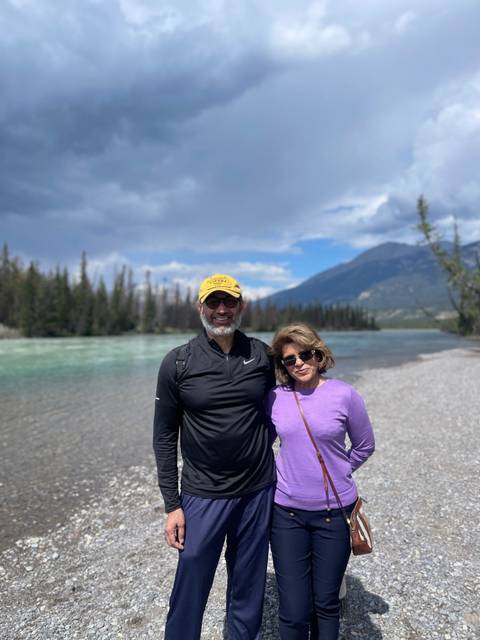 Two people smiling by a river with mountainous landscapes.