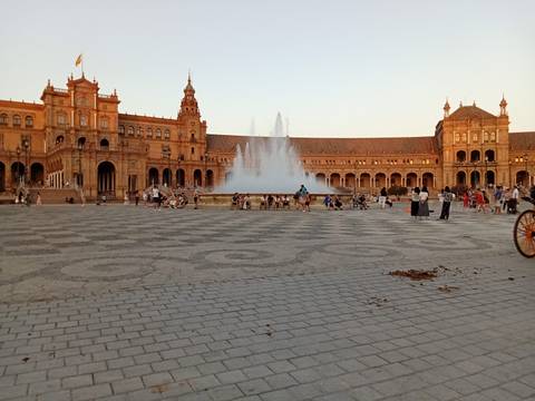       A large plaza with a fountain and a grand building, people walking around.
  
