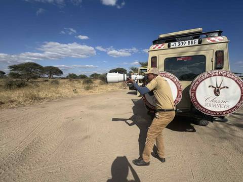 Photographer with a large lens near a safari vehicle in a savanna landscape.