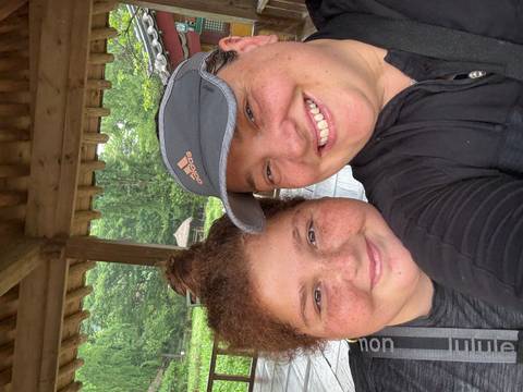 Two people smiling inside a traditional wooden structure with greenery in the background.