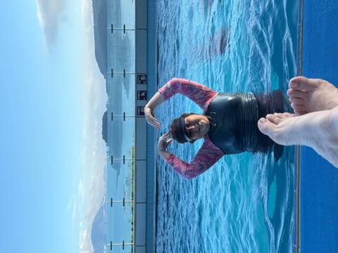 Person in a pool making a heart shape with hands, with ocean view in background.
