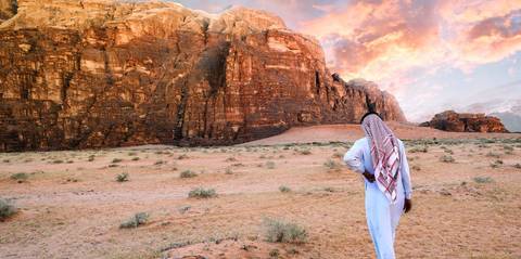       Person in traditional attire standing in front of dramatic rock formations at sunset.
  