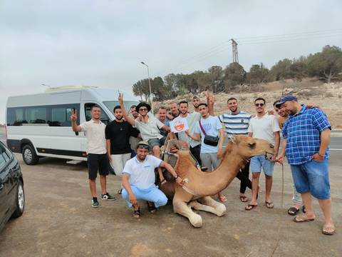       Large group of men posing with a camel by a van.
  
