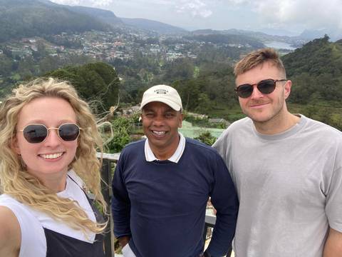 Three people smiling in front of a scenic overview of a town with greenery.
