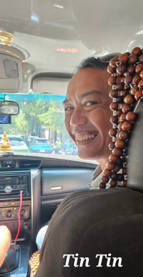 Man smiling inside a car, holding beads and a small statue on the dashboard.