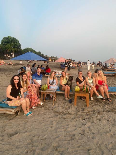 Group hanging out on the beach with drinks and a dog.
