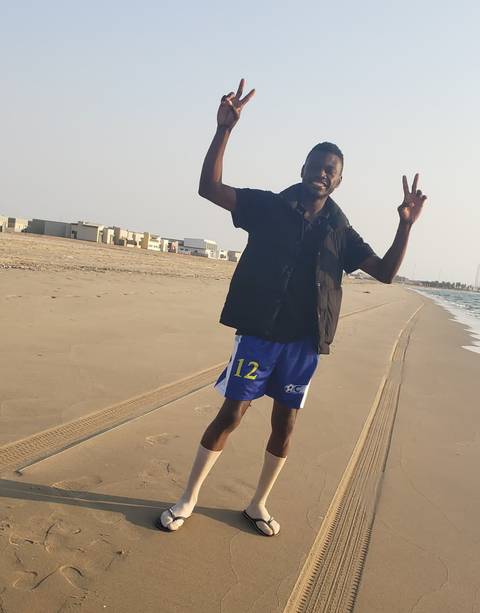 A man posing on a sandy beach with buildings in the background.