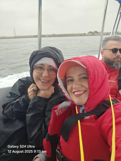       Two women smiling in rain jackets on a boat with water in the background.
  