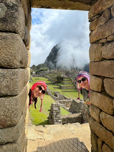       Two people peeking out from ancient ruins with Machu Picchu in the background.
  