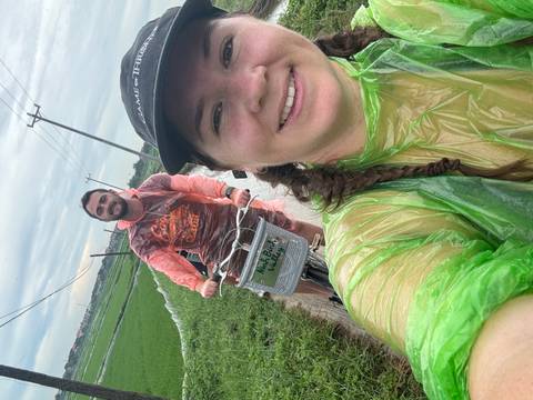       Two people cycling in rain gear along a path through rice fields.
  