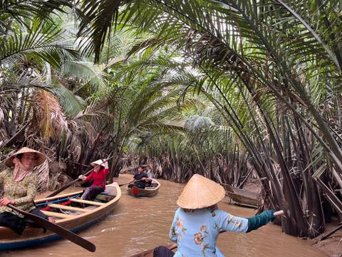 People paddling wooden boats through narrow water channels with dense vegetation.