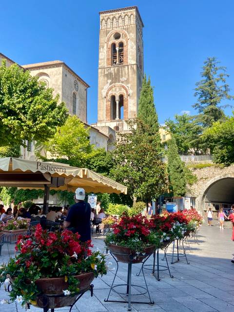 People dining outdoors under a large patio umbrella with a church tower nearby.