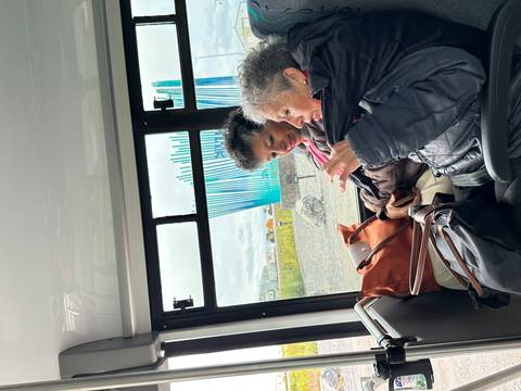       Two people seated inside a bus with outdoor scenery visible through the window.
  