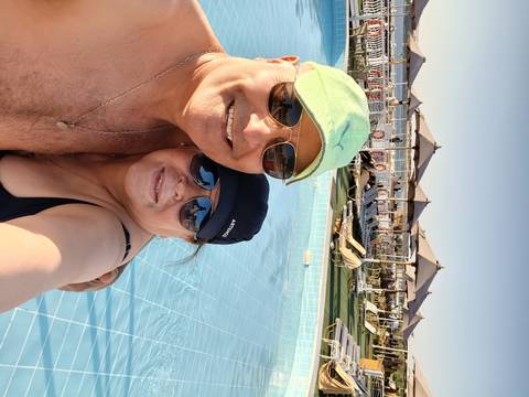 Two people taking a selfie by a swimming pool with sun loungers in the background.