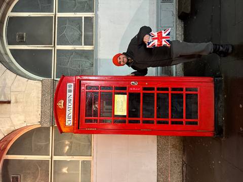 Person posing beside a red telephone booth, holding a Union Jack bag.