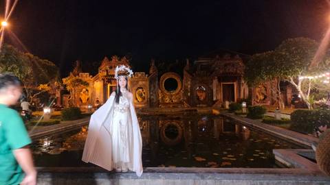 Woman in detailed costume by a pond with ancient buildings.