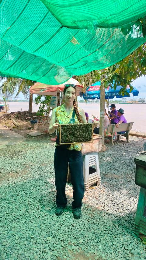 Woman holding a frame covered in bees by a river.