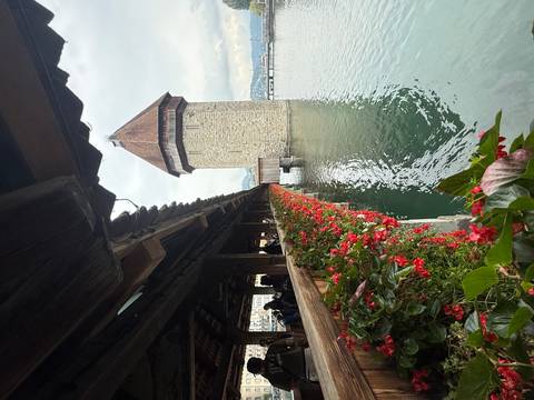 A stone tower with flowers along a covered wooden bridge over water.