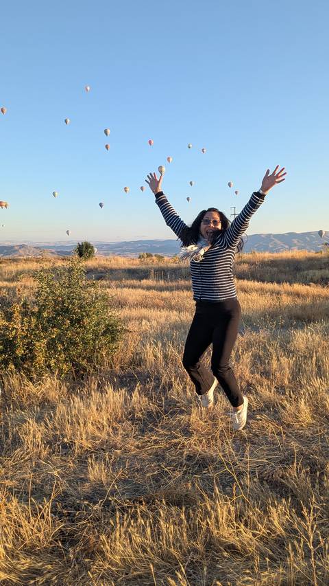       Woman joyfully jumping in a field with hot air balloons in the background.
  