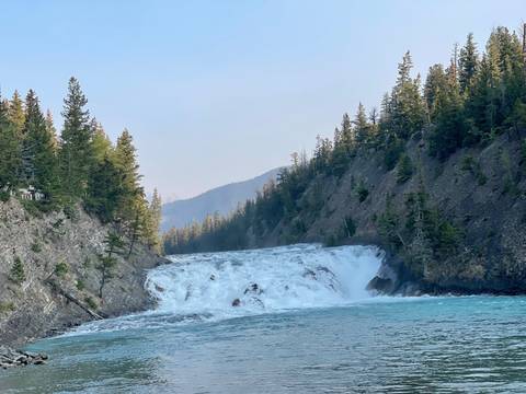 Waterfall cascading into a clear blue river flanked by cliffs and trees.