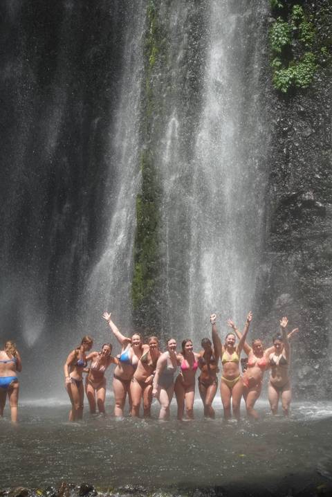       Group of people enjoying a waterfall, raising hands in joy.
  