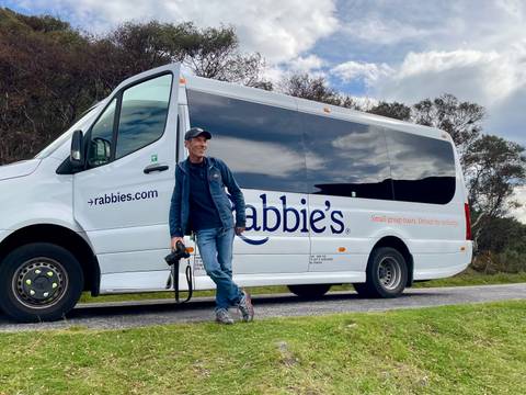 A man leaning against a tour bus with 'rabbie's' branding in a scenic outdoor setting.