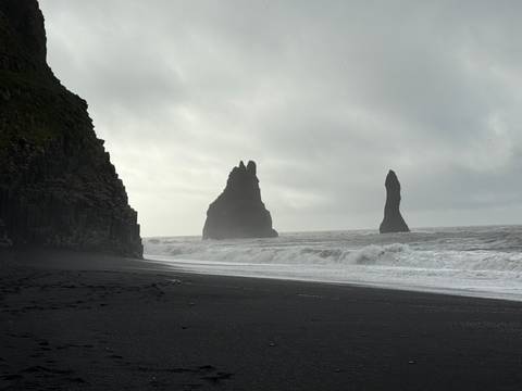       Cliffs and sea stacks under cloudy skies on a black sand beach.
  