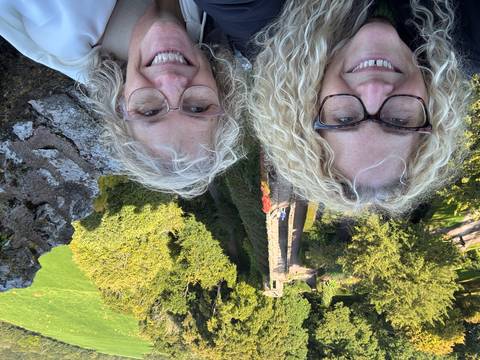 Two women smiling with a scenic background, standing on a wall.