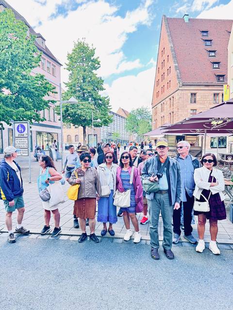 Group of people standing on a street with shops and trees.
