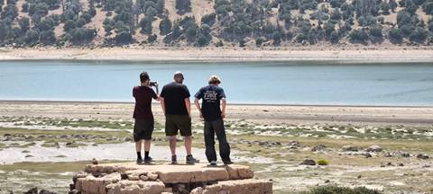 Three people standing and looking at a lake with hilly terrain.