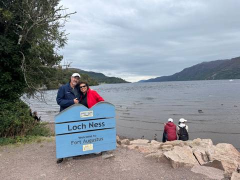 A couple posing beside a Loch Ness sign with a lake and hills in the background.
