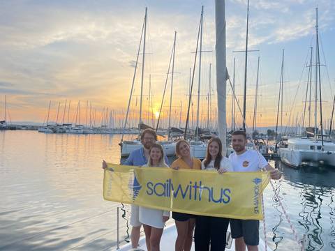 Group holding a 'sail with us' banner in front of a marina during sunset.