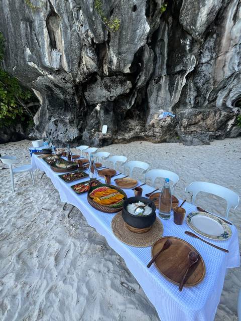 A long table set for a meal on a sandy beach next to rocky cliffs.
