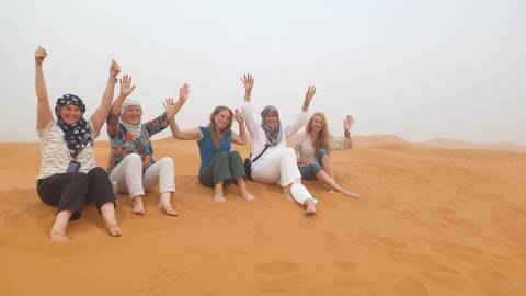       Group of people sitting on sand dunes, raising hands.
  