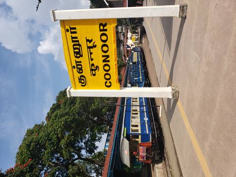 A train station with a sign for Coonoor and a blue train on the platform.