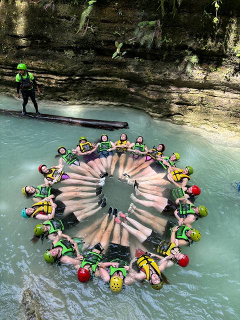 People holding hands in a circle wearing helmets and life vests in water.