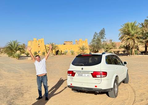 Man joyfully posing beside a SUV in front of a desert building.