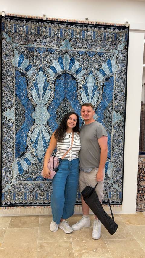      Couple posing in front of a wall-sized textile artwork.
  