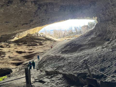 Inside a large cave with people exploring, light coming from the entrance.