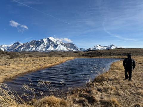 Hiker stands by a small waterbody reflecting mountains in the distance.