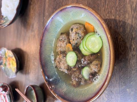       A bowl of soup with meatballs and slices of cucumber, part of a meal preparation.
  