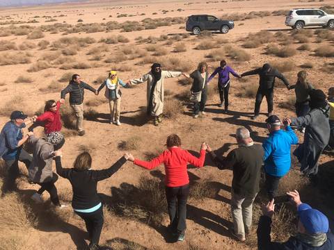       Group of people holding hands in a circle in a desert landscape.
  